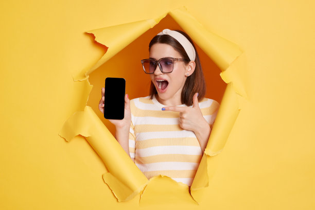 室内照片，一名穿着条纹t恤，戴着发带和墨镜的惊讶女子，从黄色墙壁上的纸洞中钻出来，指着显示为空的手机。图片下载