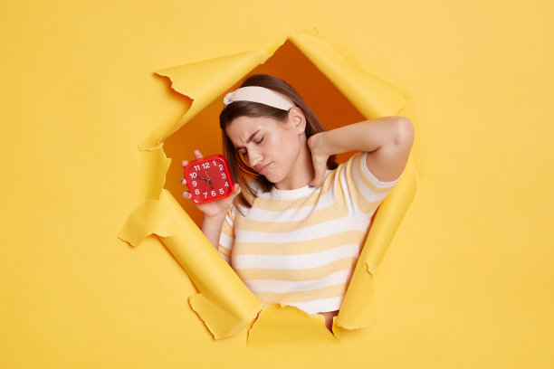 照片中穿着条纹t恤和发带的不健康精疲力竭的女子，穿过黄色墙壁的纸洞，显示闹钟，感到脖子疼痛。图片下载