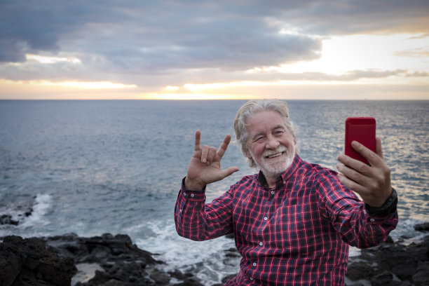 愉快的微笑着胡须的高级男子坐在海边的悬崖上，地平线上的水和日落的光。积极快乐的退休人士用手机自拍图片下载
