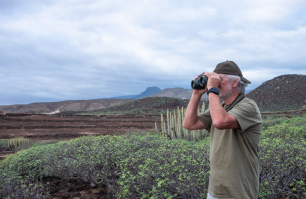 活跃的老人戴着帽子，在大自然中享受徒步旅行，站在双筒望远镜看着山景，退休老人享受假期和自由图片下载
