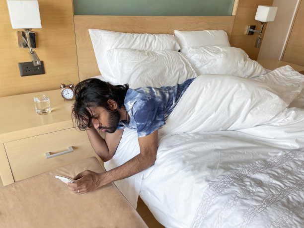 印度男子从酒店双人床的羽绒被下探出身子，查看智能手机、闹钟和床头柜上的水，专注于前景图片下载
