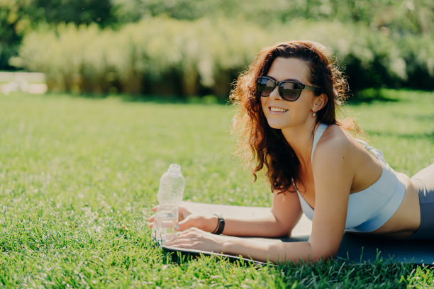 高兴放松的黑发女子的照片在健身垫上戴着时尚的太阳镜和运动服喝水，因为运动后感到口渴，躺在绿色的草地上享受美好的阳光。图片下载