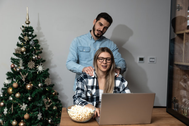 年轻夫妇自由职业者在新年或平安夜在公寓的笔记本电脑上装饰的树在网上工作。独居男女与家人朋友视频通话图片下载