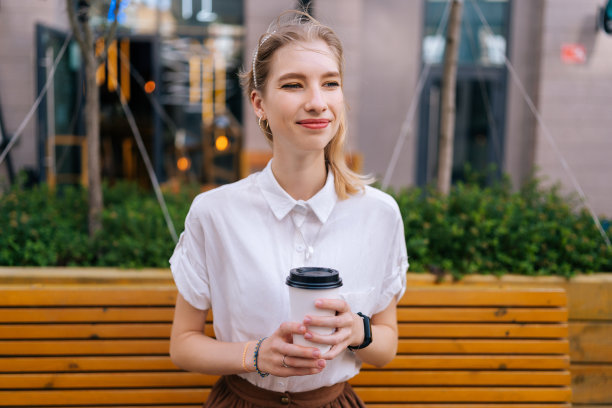 中景照片，一个快乐漂亮的年轻女子坐在黄色的长凳上，手里拿着一杯外卖咖啡，微笑着看向别处。图片下载
