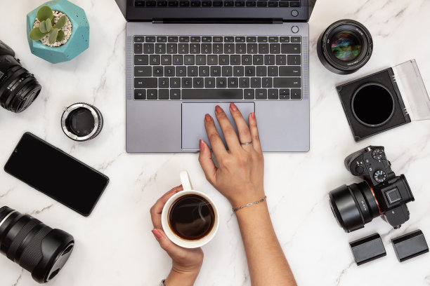 俯视图的现代办公桌的女摄影师拿着一杯咖啡与笔记本电脑，相机，手机和镜头在桌子上图片下载