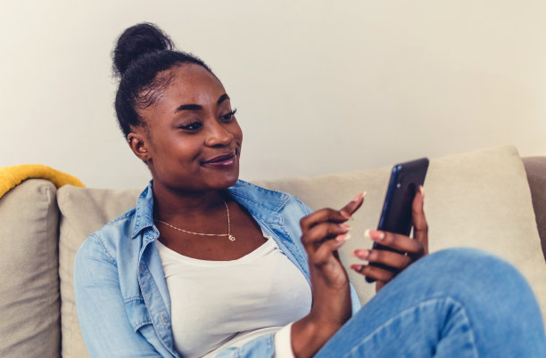 开朗的年轻女子在家里的沙发上放松时使用她的手机。快乐的微笑非裔美国妇女与智能手机坐在家里的沙发上。技术和人的概念。图片下载