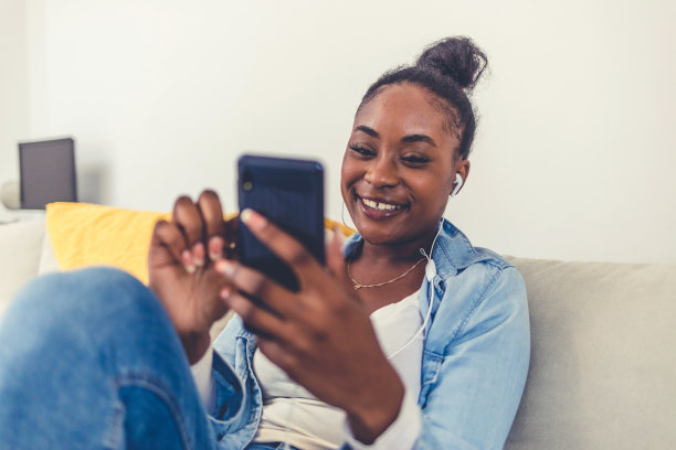 一名年轻女子在家中使用手机时戴着耳机。年轻美丽的非洲女子在休闲服装享受音乐和微笑，而在家里休息。图片下载