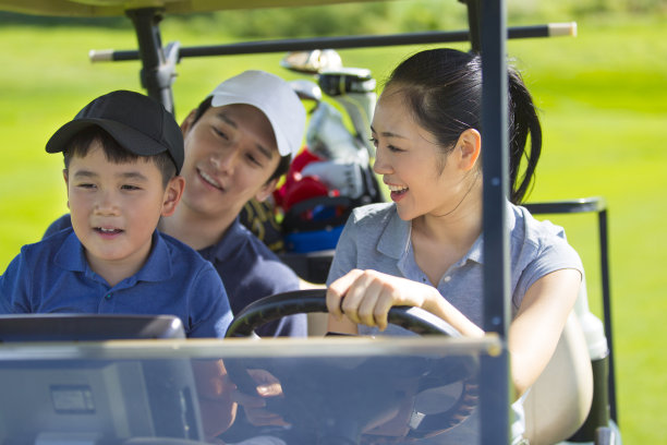 年轻的东亚三口之家在晴朗的日光下在高尔夫球场上驾驶高尔夫球车-资料照片图片下载