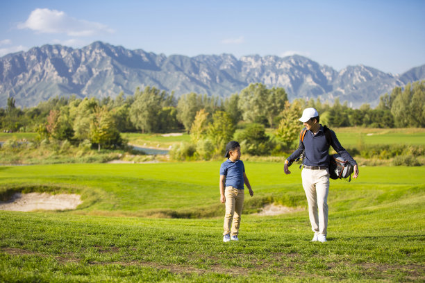 年轻的东亚父亲和儿子在阳光明媚的高尔夫球场上散步和交谈，背后有一个高尔夫球袋图片下载