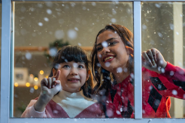 可爱的孩子看着窗外，和妈妈一起欣赏雪花。小女孩坐在家里的客厅里，看着外面的雪飘落，和家人一起庆祝圣诞节，感恩节图片下载