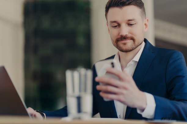 剪裁的严肃的男性企业家穿着优雅的衣服，使用现代手机和笔记本电脑，在现代办公室的室内，在模糊的背景下进行新项目的工作。图片下载