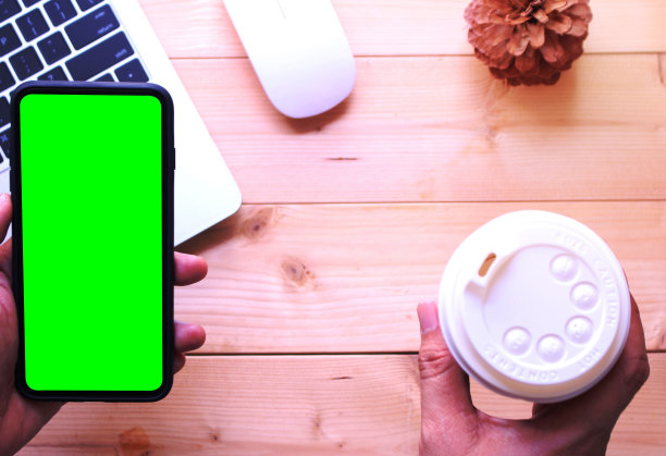 商务人士拿着绿屏智能手机，咖啡杯放在办公桌背景上图片下载