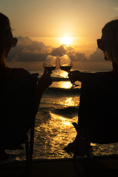 特写剪影的两个性感的年轻女子在太阳镜和比基尼坐在海边的折叠椅和碰杯葡萄酒对一个美丽迷人的日落的背景图片下载