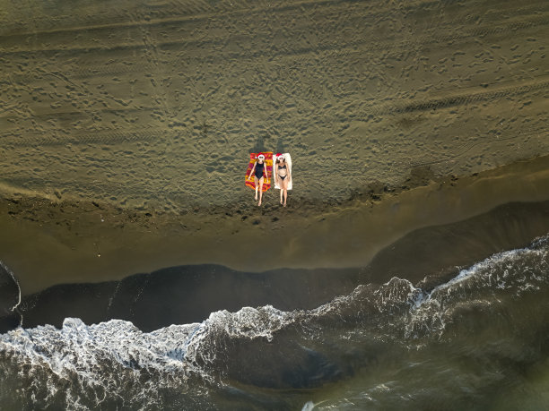 这是一架无人机从空中拍摄的照片，日落时分，两个戴着圣诞老人帽子的苗条年轻女子躺在黑海的沙滩上，海浪翻滚。海上寒假图片下载