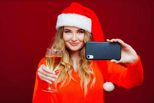 一个女人戴着圣诞老人的帽子，红色背景是香槟和智能手机。图片下载