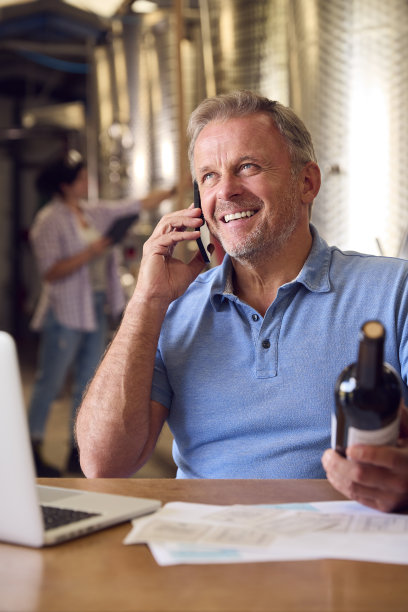 成熟男子在手机上用笔记本电脑检查酒庄内酒瓶的标签图片下载
