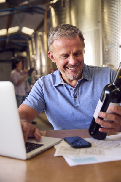 带着笔记本电脑的成熟男子在有储罐的酒庄检查瓶子上的标签图片下载