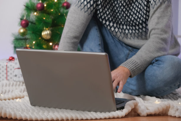 男人拿着笔记本电脑工作，远程学习2023年，圣诞节。背景圣诞树上戴圣诞帽的人，花环，礼物。远程工作。网络冲浪。男人在网上庆祝节日。图片下载