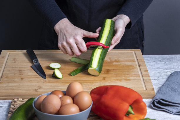 工作妇女的手在厨房木板上剥小胡瓜。前面褪色了，灰色的碗里有鸡蛋，红辣椒和另一个小胡瓜。食物准备的概念。图片下载
