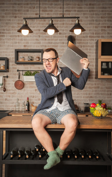 年轻愤怒的帅哥穿着西装和内裤坐在厨房桌子上砸他的笔记本电脑图片下载