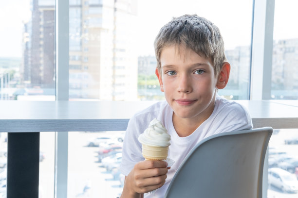 一个可爱的十几岁男孩坐在咖啡馆靠窗的桌子旁，吃着华夫饼杯里的白色冰淇淋图片下载