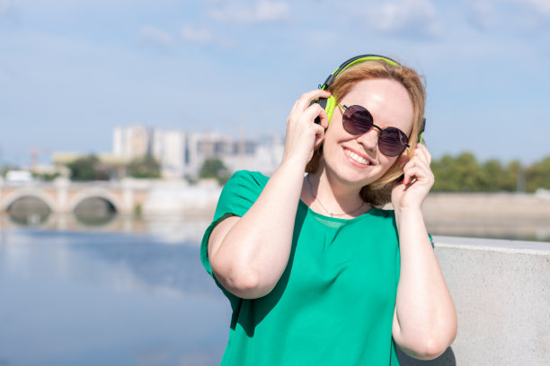 肖像一个微笑的快乐女孩戴着太阳镜和耳机，在河边听音乐。图片下载