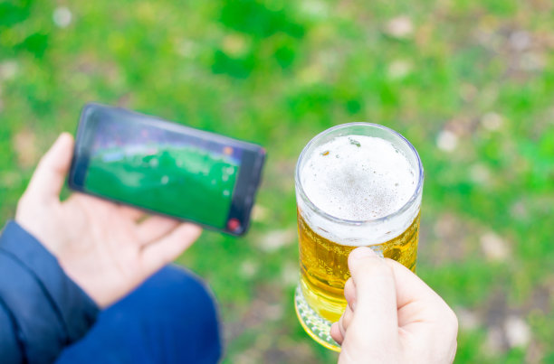 在公园里，一个年轻人正拿着一杯啤酒，用手机看足球。足球迷，世界杯。足球，偶像，忙碌。足球狂热,最喜欢的图片下载