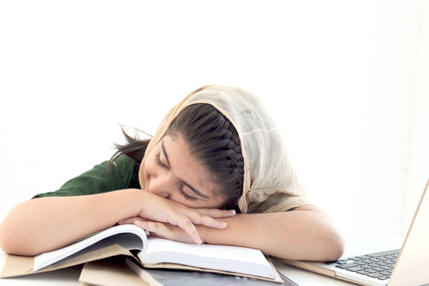 可爱的巴基斯坦穆斯林睡眼惺忪的女孩戴着头巾打盹，在桌子上学习和做作业时感到无聊和疲惫，学生孩子在白色背景上看书。图片下载