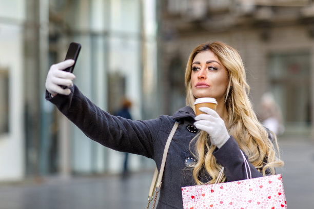 一个迷人的年轻女子站在城市的街道上，喝着咖啡，用手机自拍。图片下载