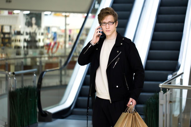 一个戴着眼镜、带着纸袋的年轻人在购物中心一边打电话一边购物图片下载