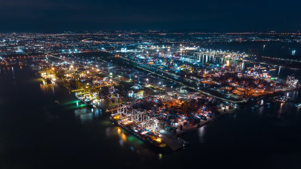 鸟瞰国际航运港口，通过起重机将集装箱装卸到运输船和拖车上，用于进出口，在照明的城市景观背景下将产品分发给客户和消费者图片下载