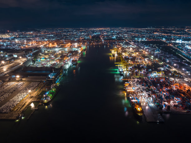 鸟瞰国际航运港口，通过起重机将集装箱装卸到运输船和拖车上，用于进出口，在照明的城市景观背景下将产品分发给客户和消费者图片下载