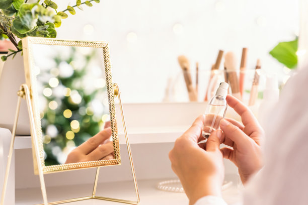 女人用香水在她的手上，坐在化妆台附近的配件和桌子镜子。圣诞节，灯泡，桉树，冷杉，松枝插在花瓶里。图片下载