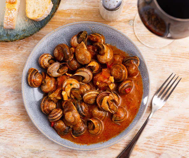 红酱炖蜗牛放在灰色的陶瓷盘子里，靠近餐厅里的餐具和面包图片下载