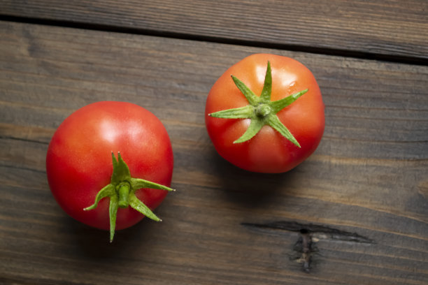 木桌上放着两个红西红柿和绿尾巴。红番茄是一种健康的食物，有明亮的花梗。木制背景上有两个成熟的西红柿。图片下载