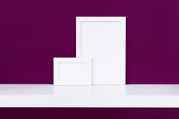 在明亮的紫红色墙壁背景的白色架子上，有两个白色的木制空相框。空白纸框架，现代家居装饰模型。图片下载