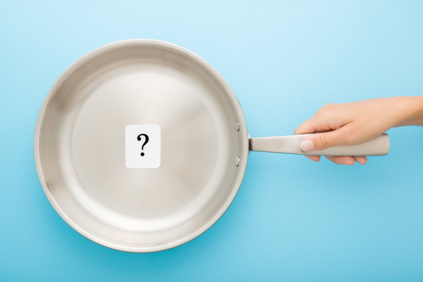 年轻的成年女子手握不锈钢煎锅，浅蓝色的桌子背景上有黑色问号的白色卡片。特写镜头。柔和的颜色。烹饪概念。自顶向下视图。图片下载