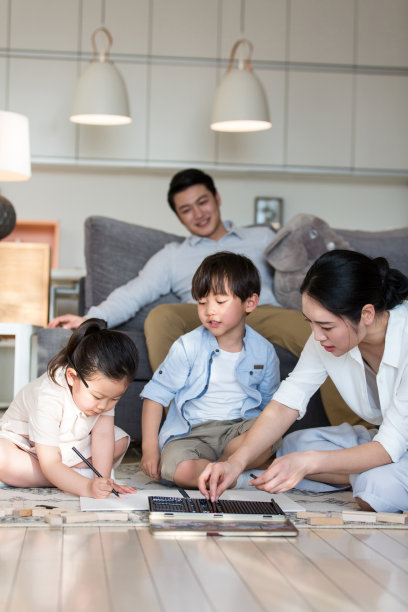 年轻的妈妈耐心地坐在地毯上和孩子们用铅笔画，而爸爸则舒服地坐在沙发上——stock photo图片下载