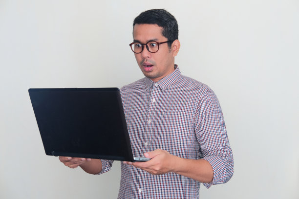 成年亚洲男子看着他的笔记本电脑屏幕，表情震惊图片下载