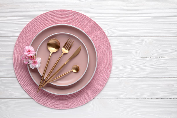 粉红色的陶器和金色的花餐具，白色的木桌上摆着静物。前视图。上菜和吃饭用的盘子和用具。现代陶瓷圆边板。副本的空间。图片下载