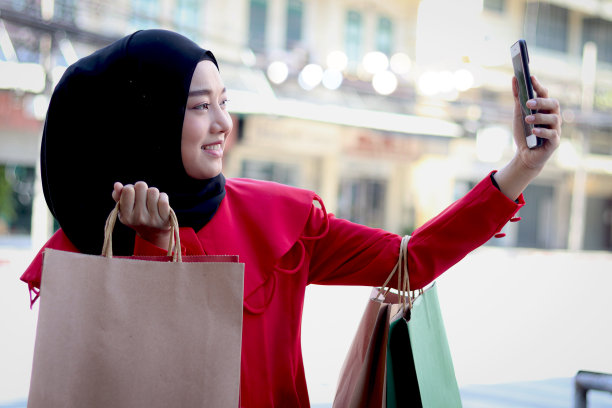 快乐的微笑年轻的亚洲穆斯林妇女戴着头巾，穿着红色连衣裙，拿着手机自拍，走在步行街上，美丽的购物肖像走在城市道路上。图片下载