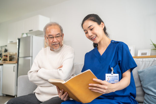 亚洲女性护理员在疗养院照顾老年男性。年轻的女治疗师医生坐在沙发上，读着快乐的书给年长成熟的男病人。医疗保险服务理念图片下载