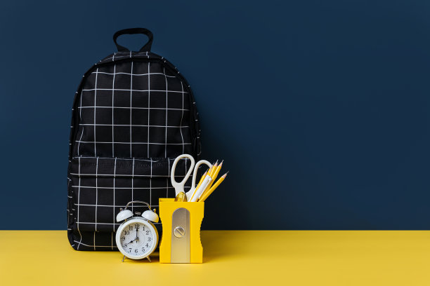 黑色格子双肩包与学校文具在黄色和蓝色的背景。办公用品和书包在桌子上。概念回到学校。图片下载