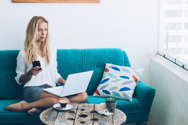 沉思的女性IT专业人员琢磨着笔记本电脑系统的信息，在舒适的家庭公寓远程工作，沉思的金发女人手里拿着智能手机小工具坐在舒适的沙发上图片下载