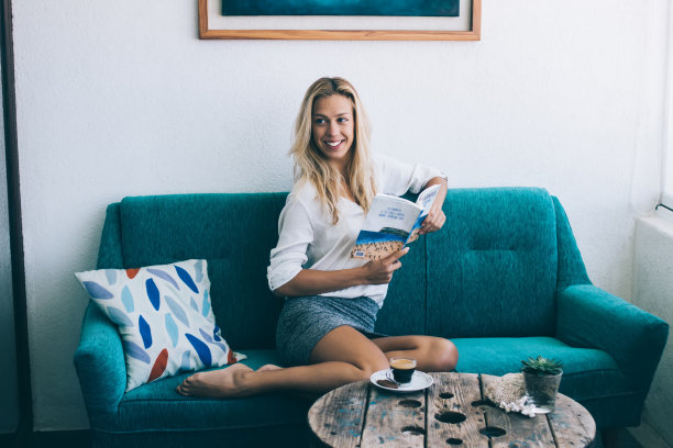 快乐的女性在舒适的家庭内部的自由周末与文学爱好，微笑的白人女性与有趣的小说书休息在舒适的沙发上靠近咖啡桌图片下载