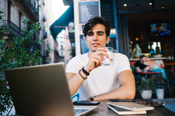 英俊的白人潮人数字游牧民的肖像喝水在咖啡馆露台上工作，积极的20多岁的男性博主拿着杯子和饮料坐在笔记本电脑远程工作图片下载