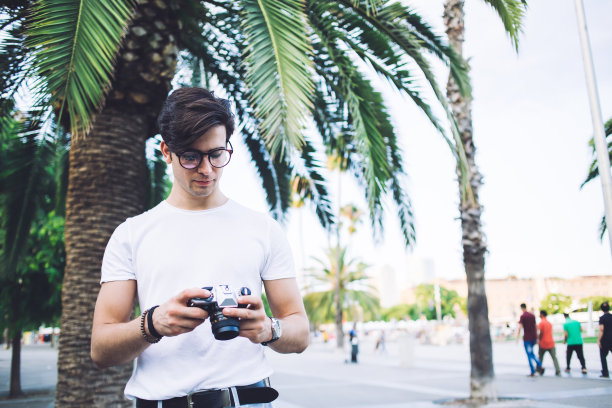 沉思的白人男性戴着眼镜，拿着相机检查照片，在度假旅行中探索小镇，熟练的摄影师站在城市广场上，棕榈树享受夏天的周末图片下载
