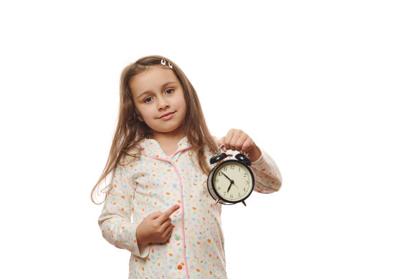 小女孩穿着舒适柔软的睡衣，指着白色背景上的黑色闹钟。睡觉时间到了图片下载