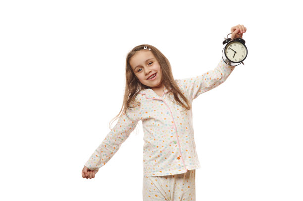 小女孩穿着舒适柔软的睡衣，抱着黑色闹钟醒来，孤立在白色背景上图片下载