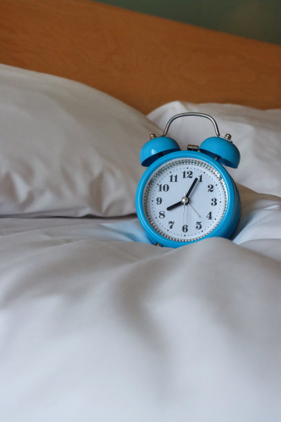 蓝色金属外壳，复古闹钟，双钟，白色表盘的模拟显示时钟坐在白色羽绒被床罩上，黑色数字，小时和分针，枕头倚在木头和玻璃床头板上，专注于前景，复制空间图片下载